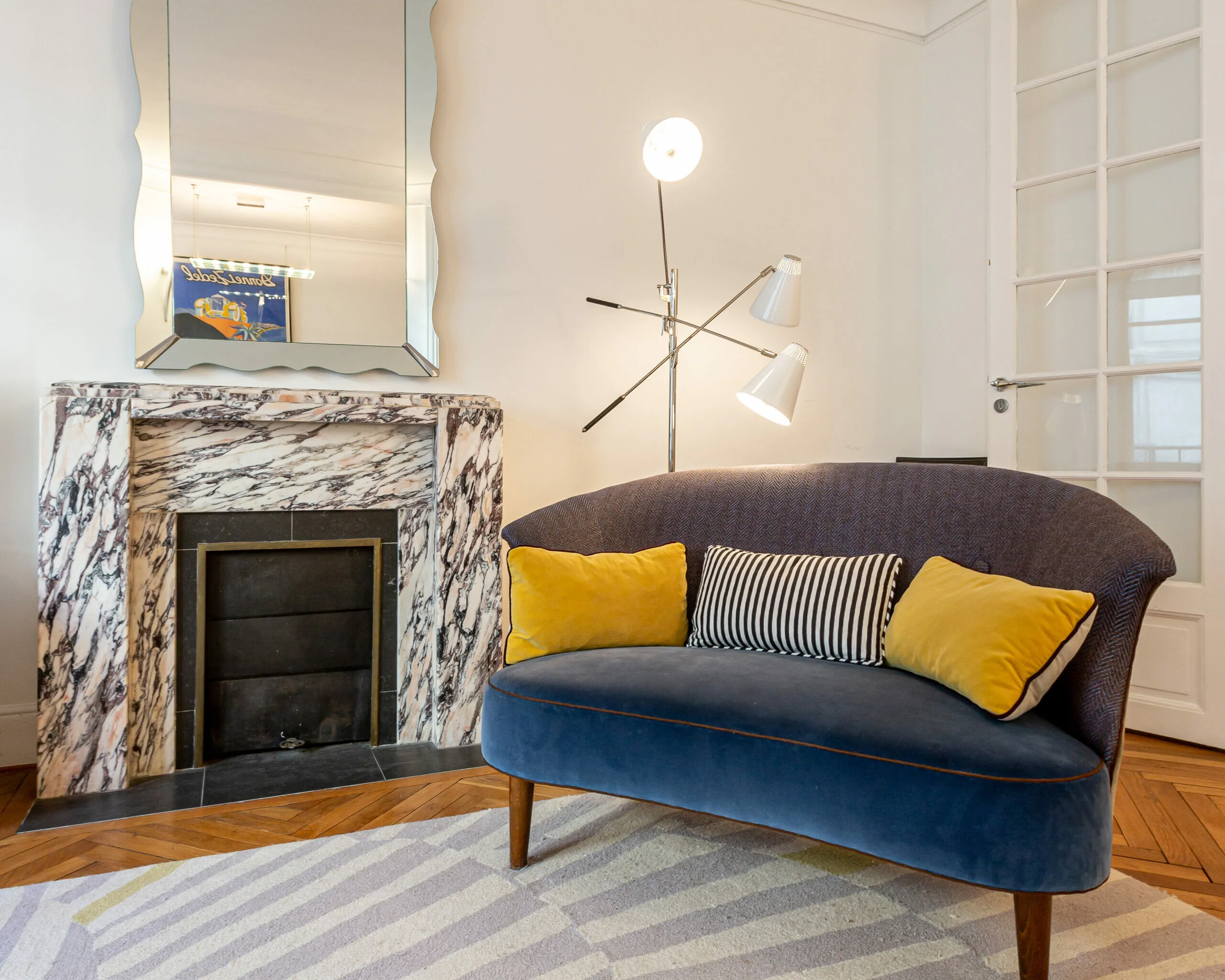 A cozy living room features a blue velvet loveseat with yellow and striped cushions, home staging accents, a modern floor lamp, a marble fireplace with a mirror above it, and a patterned rug on a herringbone wood floor.