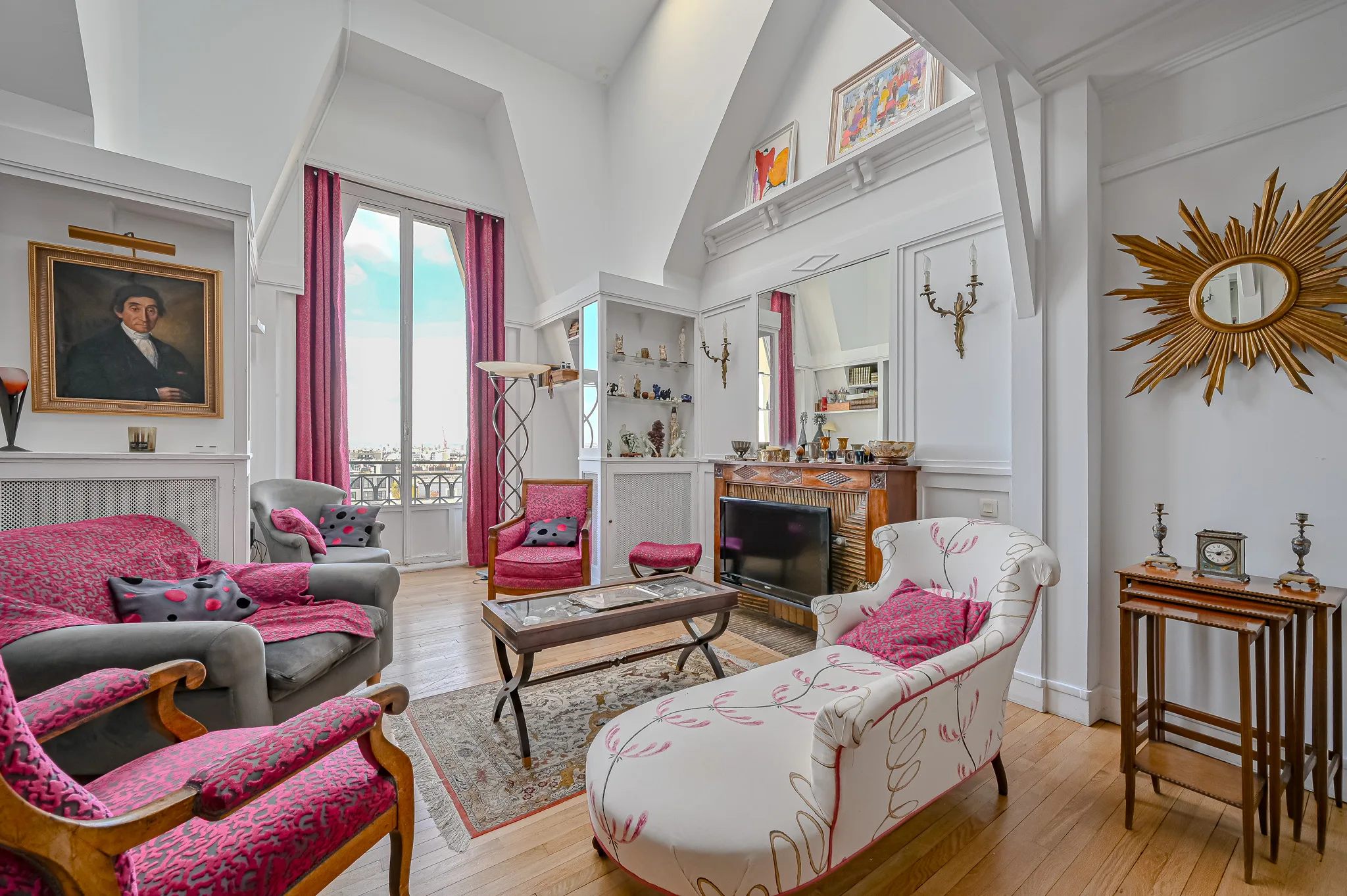 A bright and elegant living room with high ceilings, pink and white armchairs, a patterned sofa, hardwood floors, a fireplace topped with a mirror, striking artwork, large windows with red curtains, and elegant decorative accents.