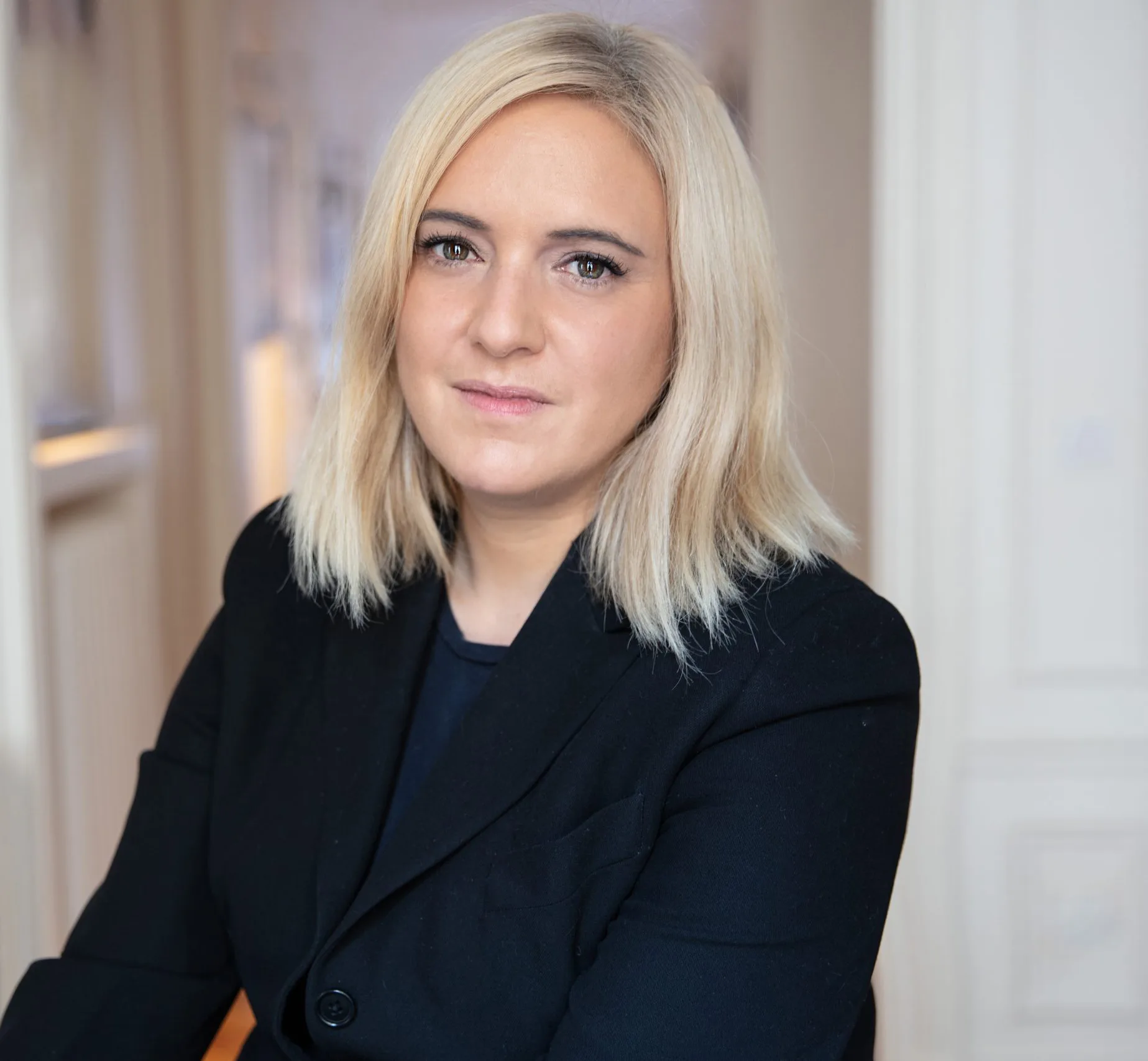 A woman with straight, shoulder-length blonde hair, wearing a black blazer, is sitting indoors, looking at the camera with a neutral expression. Her composed gaze reflects the professionalism of a luxury real estate agency. The background is slightly blurred.