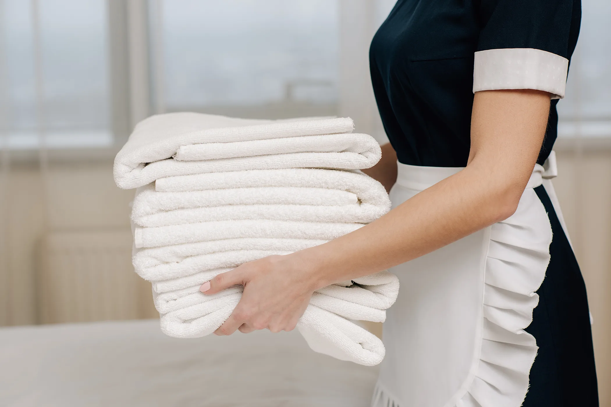 A person wearing a dark dress and a white apron, who works for an Airbnb concierge service in Paris, is standing inside near a window, holding a stack of neatly folded white towels.